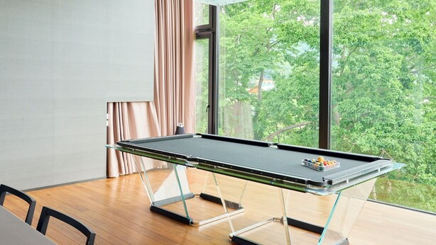 Una mesa de billar con superficie de vidrio y patas, una ventana con vista a árboles, y una mesa de madera.