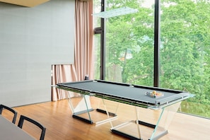 A pool table with a glass top and legs, a window with a view of trees, and a wooden table. - Nero (Abuta)