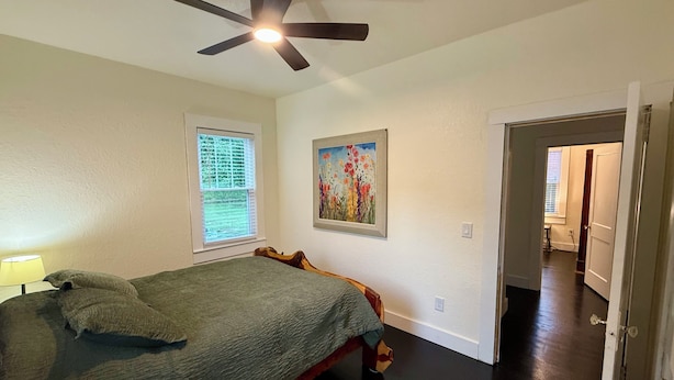 Une chambre à coucher avec un lit, un ventilateur de plafond, une fenêtre avec des stores et un tableau au mur.