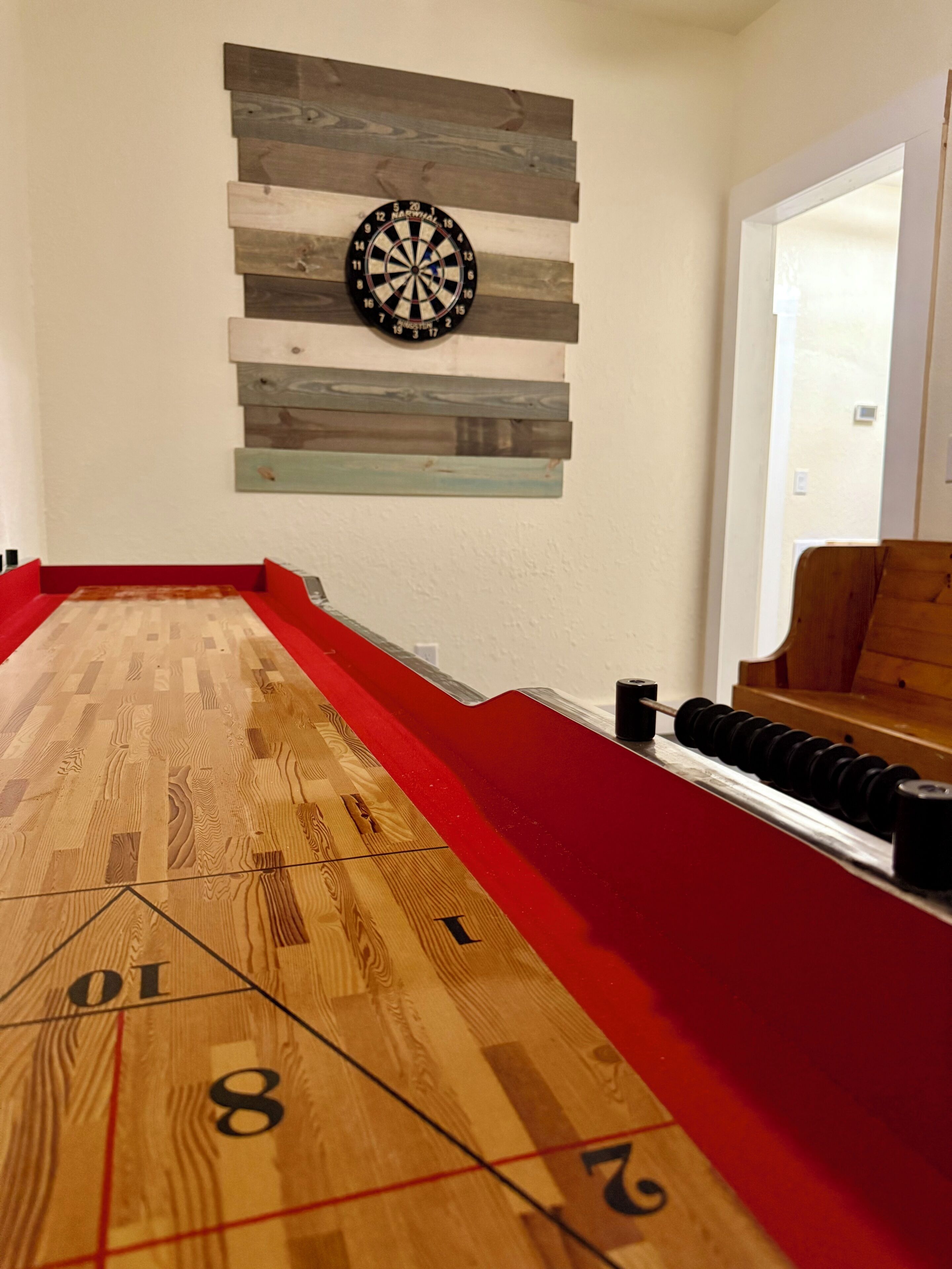 Une table de jeu de palet, avec un jeu de fléchettes fixé au-dessus, un plancher en bois et une zone recouverte d’une moquette rouge.