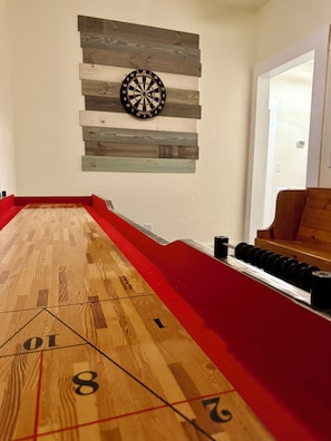 Une table de jeu de palet, avec un jeu de fléchettes fixé au-dessus, un plancher en bois et une zone recouverte d’une moquette rouge.