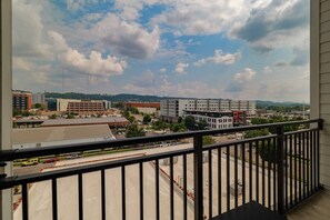 Blick von einem Balkon mit Metallgeländer auf einen Parkplatz und Gebäude, strahlend blauer Himmel darüber.