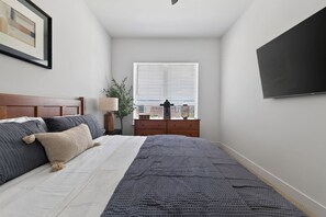 Ein Schlafzimmer mit Bett, Kommode, Lampe, einem Fenster mit Jalousien und einem Fernseher an der Wand.