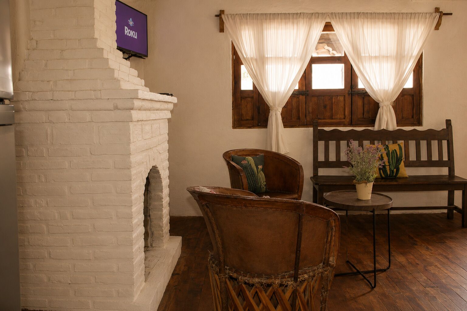 A room with a brick fireplace, a television mounted on the wall, a wooden bench, two wicker chairs, and a small table with a potted plant.