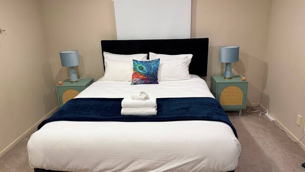 A neatly made bed with a white and blue bedspread, a decorative pillow, and a folded towel on the bed. There are two bedside tables with lamps, and a wall-mounted light fixture above the bed. - Stay Local, Live Like a Kiwi: Bright & Spacious 3BR Green Home (New Lynn)