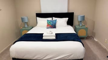 Een net opgemaakt bed met een wit- en blauwe spreidek, een decoratief kussen en een opgevouwen handdoek. Er staan twee nachtkastjes met lampen bij het bed, en een wandlamp erboven.