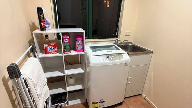 A compact kitchen with a washing machine, a small sink, and a window with a view. - Stay Local, Live Like a Kiwi: Bright & Spacious 3BR Green Home (New Lynn)