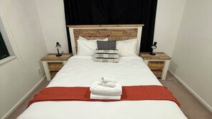 A neatly made bed with white linens and a red blanket, flanked by two bedside tables with lamps, in a room with white walls and a black curtain. - Stay Local, Live Like a Kiwi: Bright & Spacious 3BR Green Home (New Lynn)