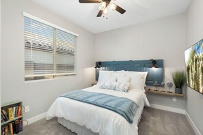 A bedroom with a bed, a ceiling fan, a window with blinds, a bedside table, a wall-mounted shelf with decorative items, and a bookshelf. - Desert Mountain Views - Quiet Retreat - Hot Tub - Firepit - Quiet Neighborhood (Goodyear)