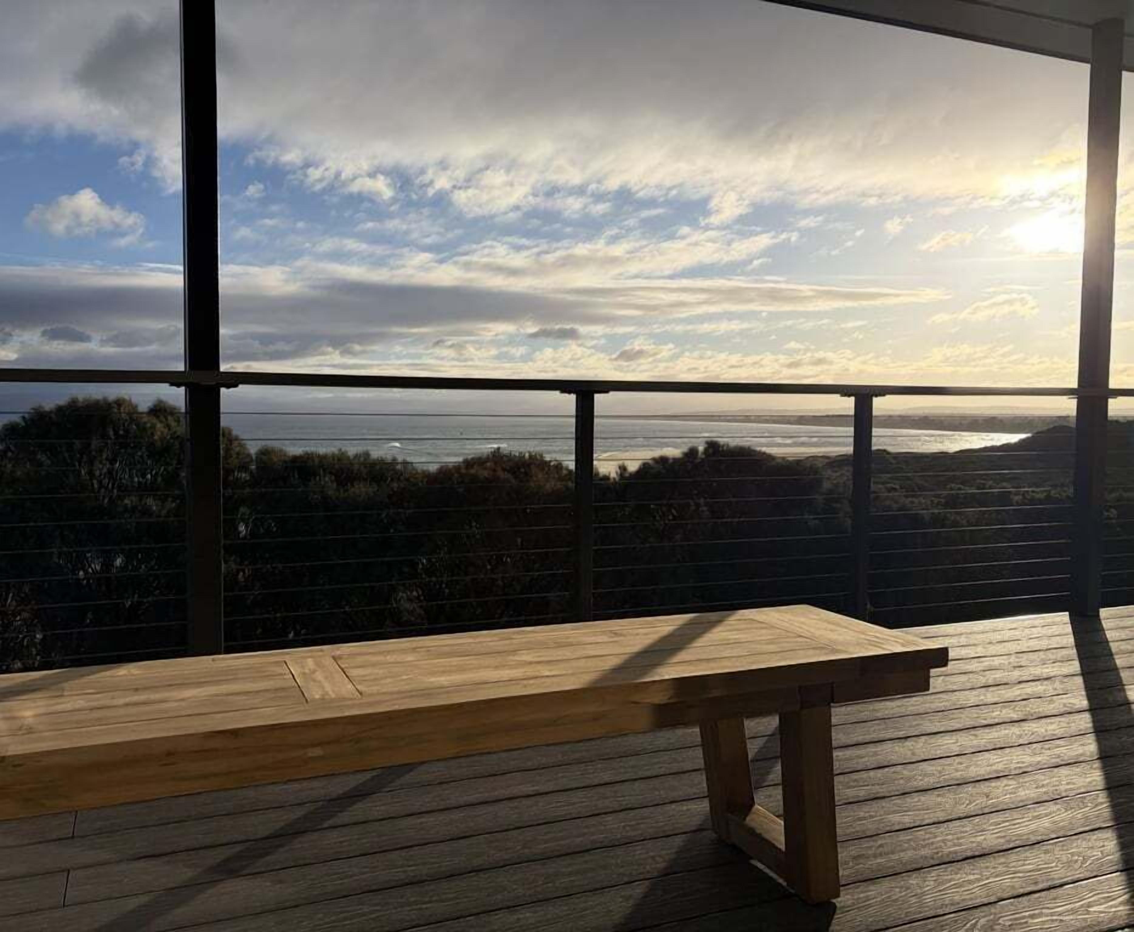 A wooden deck with a bench, overlooking a coastal landscape with trees and a body of water.