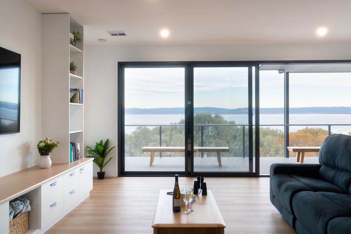 Ein modernes Wohnzimmer mit einem großen Fenster, einem Sofa, einem Couchtisch mit Flasche und Gläsern sowie einem Bücherregal.