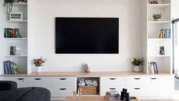 A modern living room with a flat-screen TV mounted on the wall, a bookshelf with books and decorative items, a coffee table with glasses and books, and a sofa.