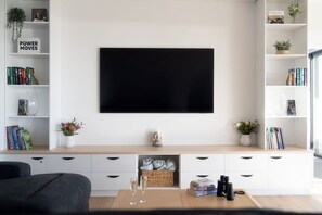 A modern living room with a flat-screen TV mounted on the wall, a bookshelf with books and decorative items, a coffee table with glasses and books, and a sofa. - Beach Break - Freycinet Holiday Houses (Coles Bay)