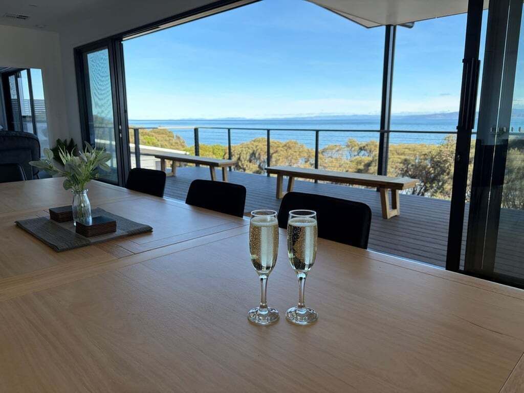 Et spisebord med to glas champagne, en vase med planter og udsigt til havet gennem store vinduer.