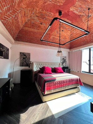 Ein Schlafzimmer mit Ziegeldecke, einem Bett mit rotem und beigem Bettbezug und einem großen Fenster mit Vorhängen.