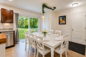 Zona pranzo con tavolo bianco imbandito per sei persone, cucina con lavastoviglie e vista su un giardino verde attraverso una porta vetrata.