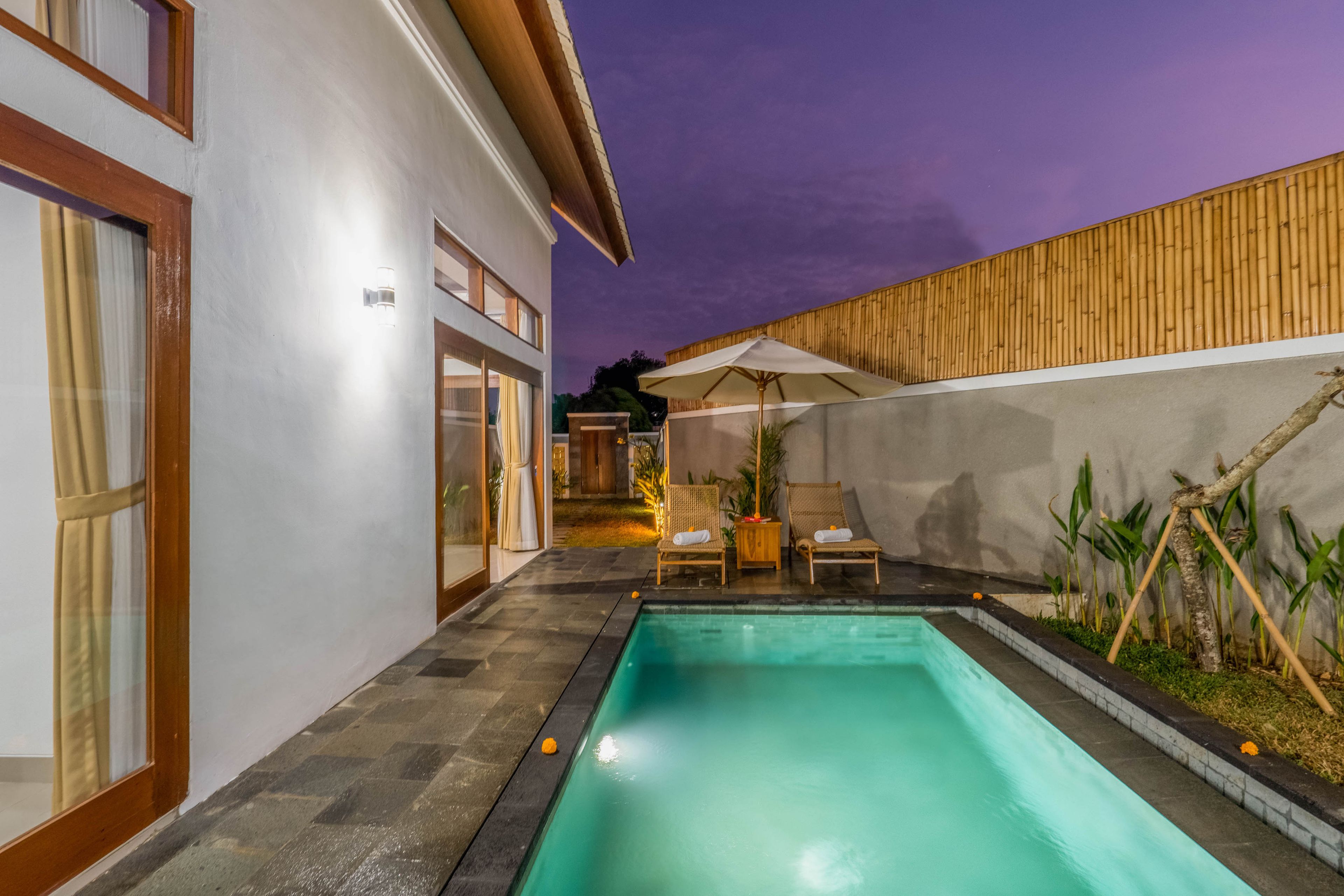 A swimming pool with lounge chairs and umbrellas, surrounded by a bamboo fence and a white building.