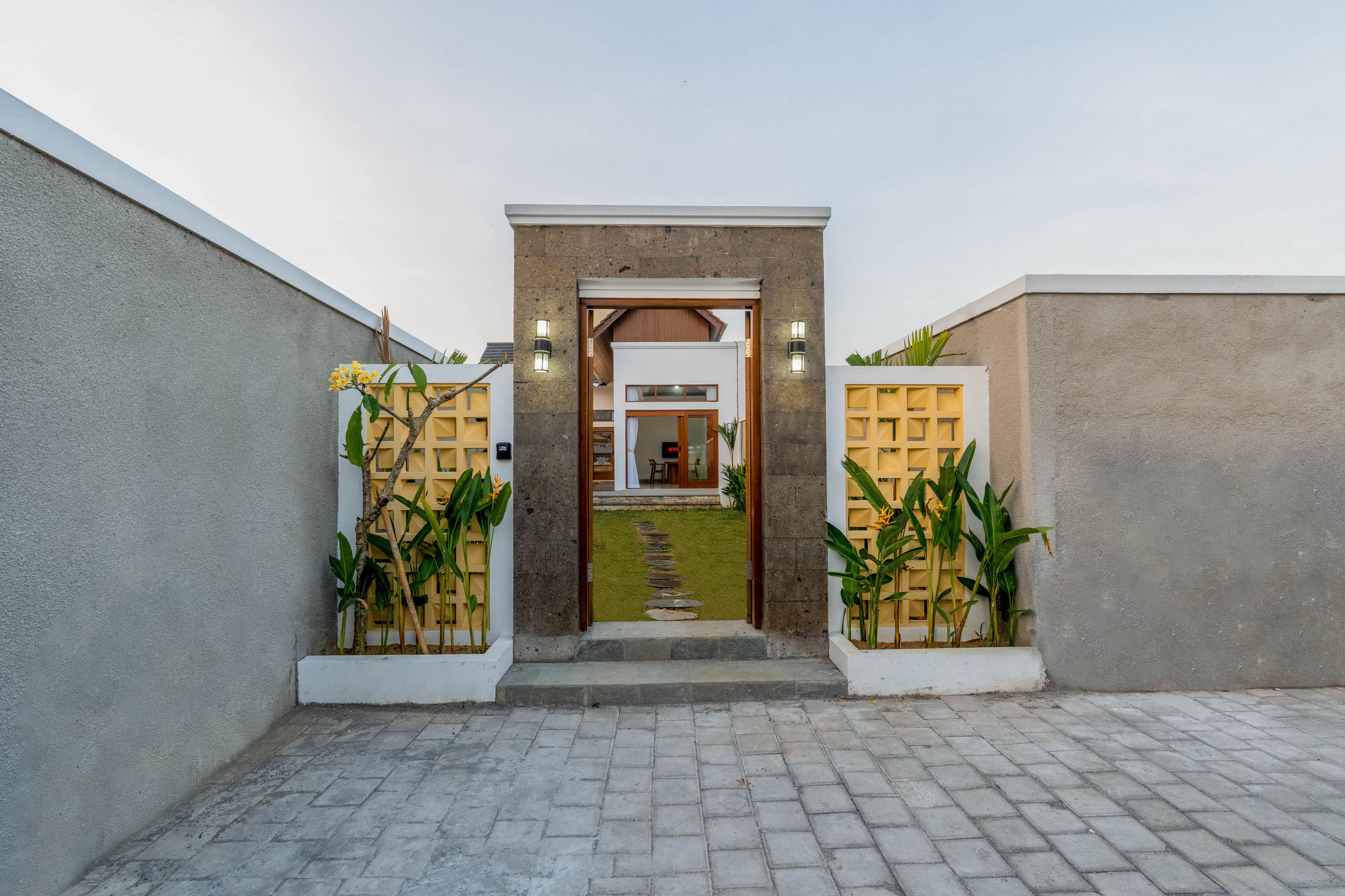 A modern entrance with a concrete pathway leading to a door, flanked by two lighted wall sconces and potted plants.