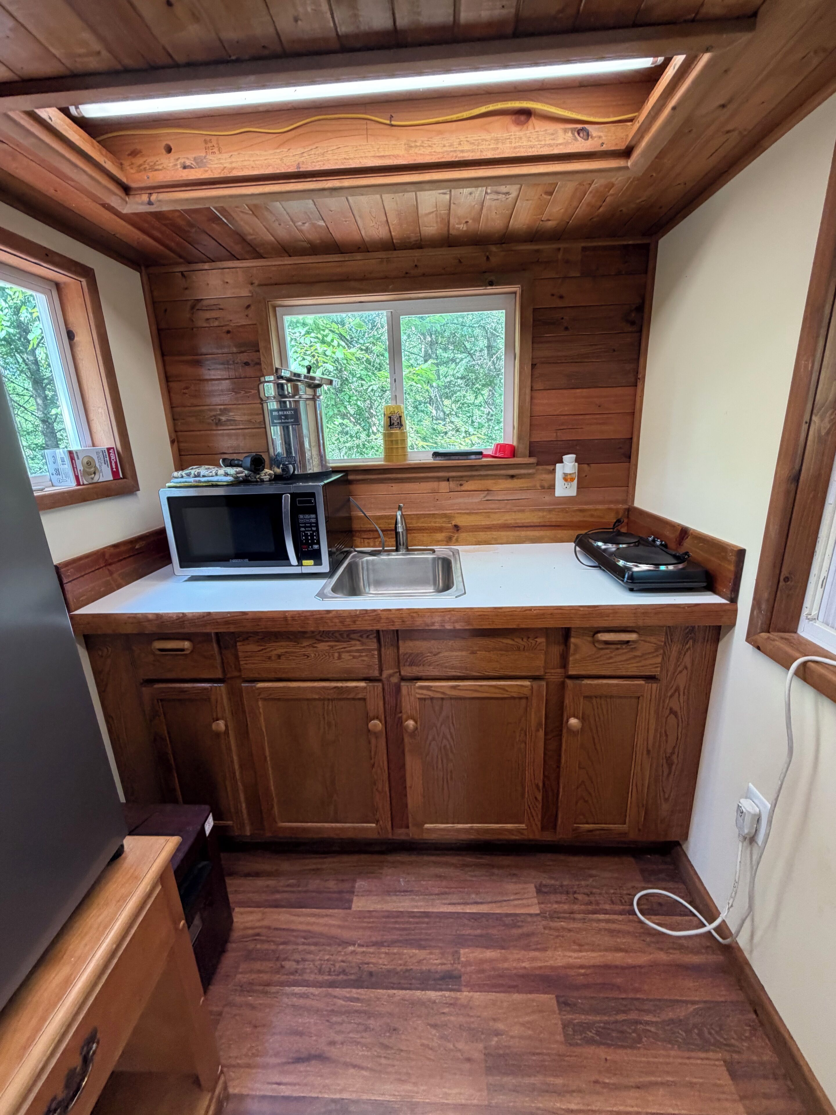 Une cuisine compacte avec des meubles en bois, un four à micro-ondes, un évier et une fenêtre donnant sur un espace verdoyant.
