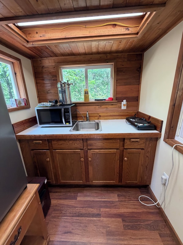 A compact kitchen with wooden cabinets, a microwave, a sink, and a window overlooking greenery.
