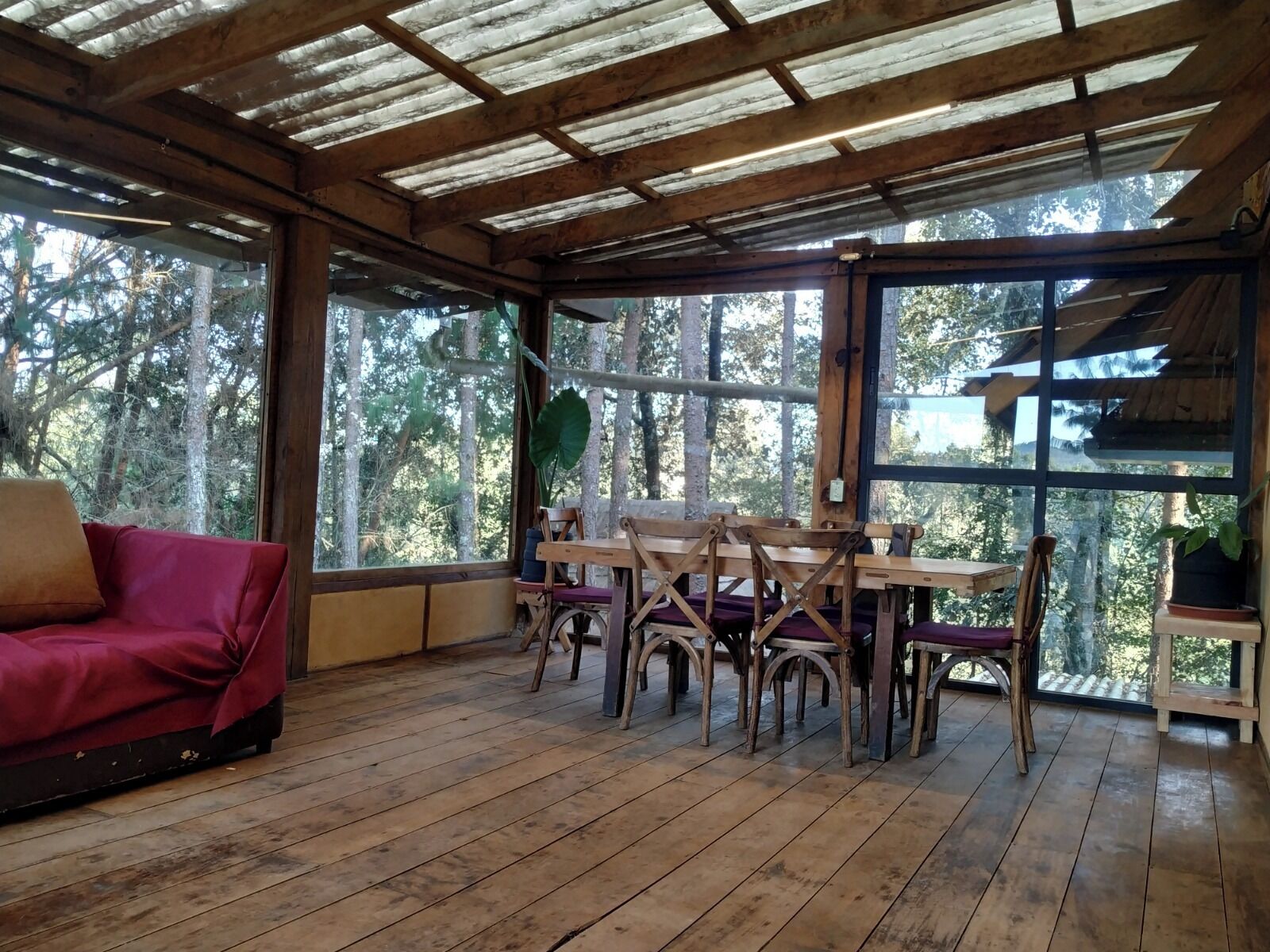A wooden interior with a red sofa, a wooden dining table with chairs, and large windows offering a view of trees.