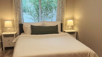 Uma cama bem arrumada com roupa de cama branca e um travesseiro verde escuro, com duas mesas de cabeceira com luminárias, num quarto com janela e cortinas.