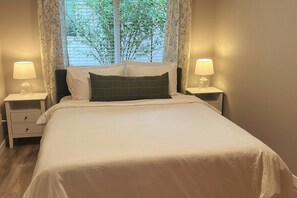 Una cama bien hecha con ropa de cama blanca y un almohadón verde oscuro, flanqueada por dos mesitas de noche con lámparas, en una habitación con ventana y cortinas.