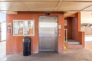 A building entrance with a metal door, a fire extinguisher, a trash bin, and a notice board with various papers. - Buena Vista 405 | Beachfront | Beach Side Pool | Low-Density | My Beach Getaways (Gulf Shores)
