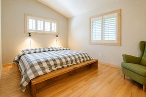 Ein Schlafzimmer mit einem hölzernen Bett, zwei Jalousienfenstern, einem grünen Sessel und einer Wandleuchte.