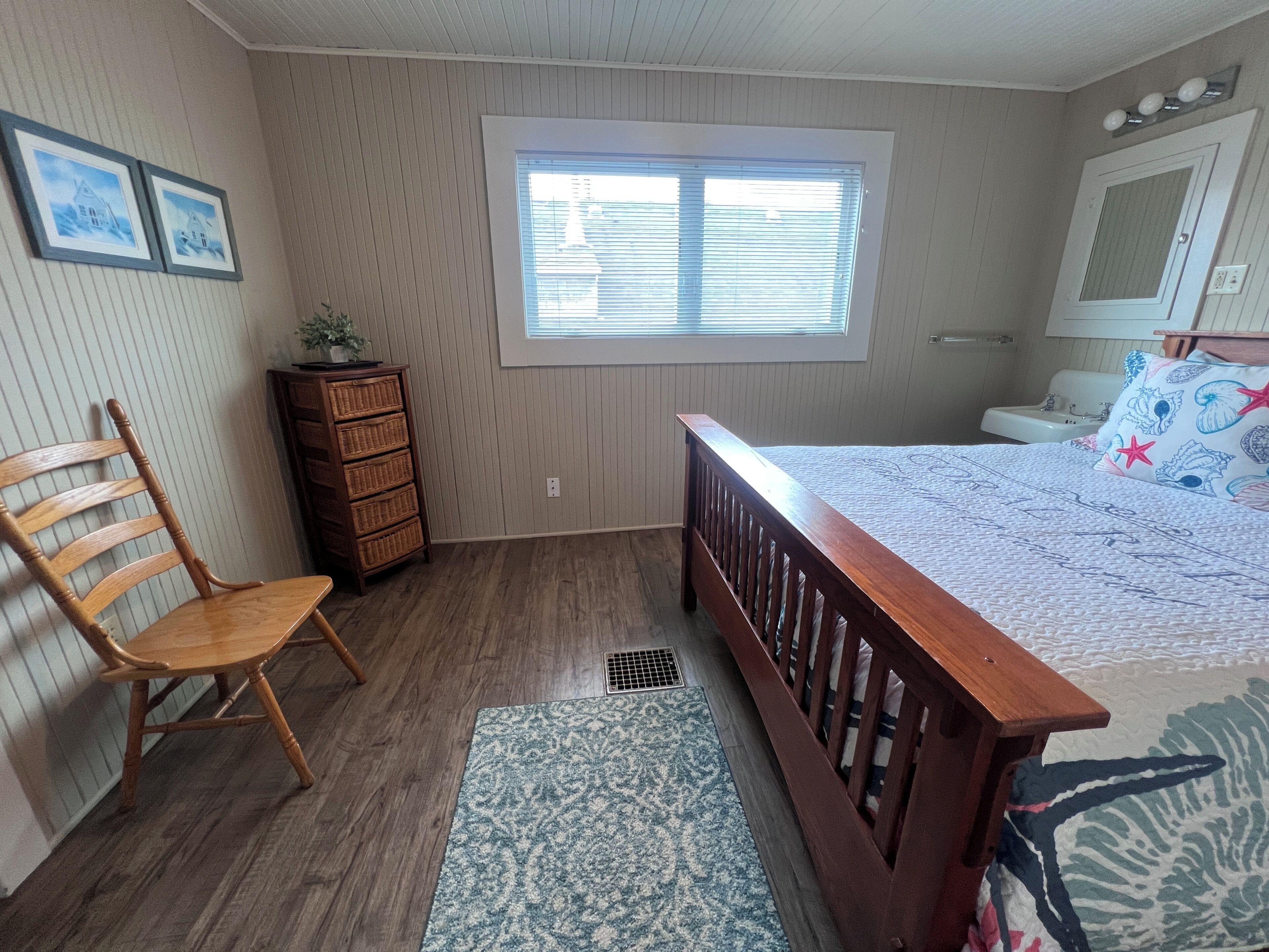 A bedroom with a wooden bed, a chair, a dresser, a window, and a bathroom with a sink and mirror.