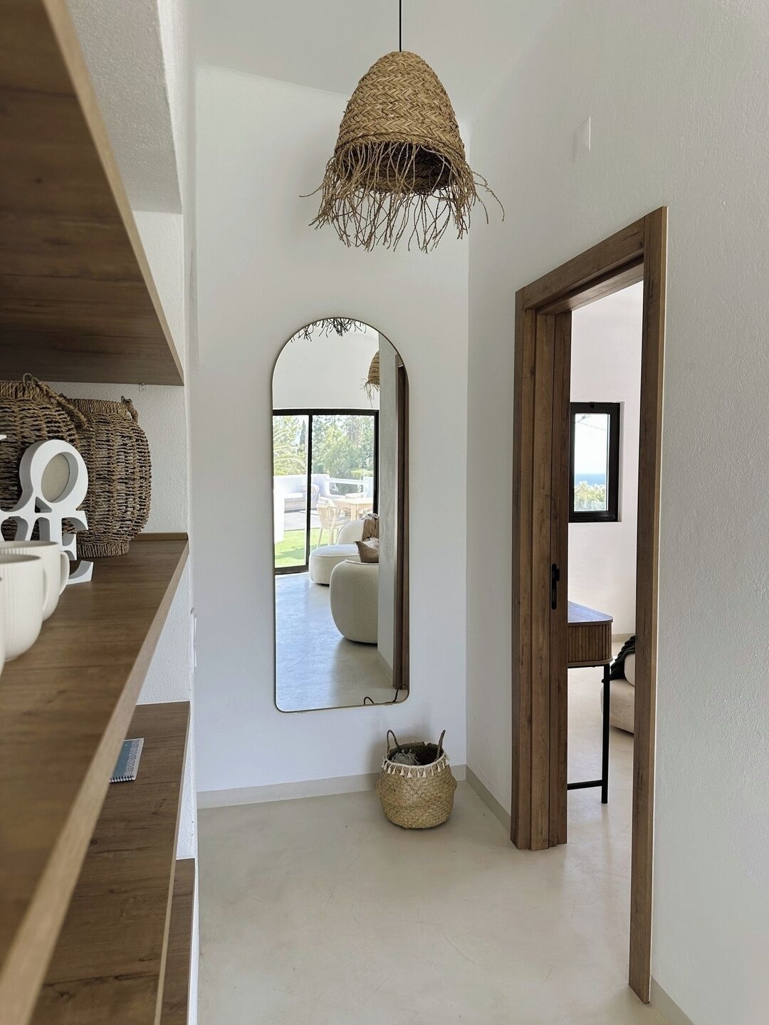 Un intérieur moderne agrémenté d’un luminaire suspendu en fibres tressées, d’un grand miroir reflétant une pièce avec un canapé et une fenêtre, et d’une porte en bois donnant sur un autre espace.