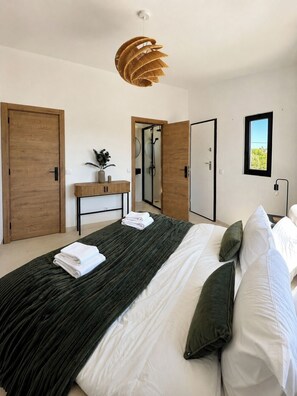 A modern bedroom with a large bed, a wooden door, a vanity, and a shower area. - Bright 2-BR Apt with Spacious Terrace and Sea Views (Carvoeiro)