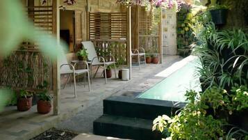 Un área de estar al aire libre con techo, sillas y mesas, rodeada de plantas en macetas y una pequeña piscina.