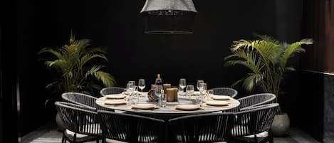 A round dining table set for six with black wicker chairs, a woven pendant light, and potted plants on either side.