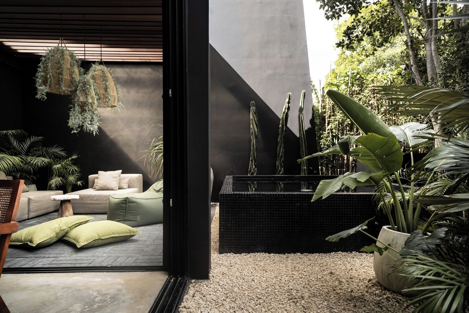An indoor-outdoor living area with a modern sofa, green cushions, hanging plants, and a black tiled water feature.