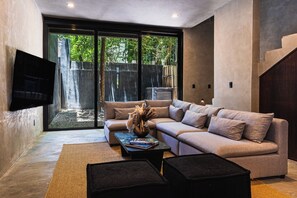 A modern living room with a sectional sofa, a coffee table with books, a flat-screen TV mounted on the wall, and a large sliding glass door leading to an outdoor area. - Casa Naya - 2 Pools, Near Beach, VIP Concierge (Tulum)