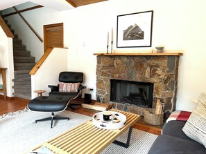 Ein Wohnzimmer mit Kamin aus Stein, einem schwarzen Ledersessel, einem Holztisch und einer Treppe.