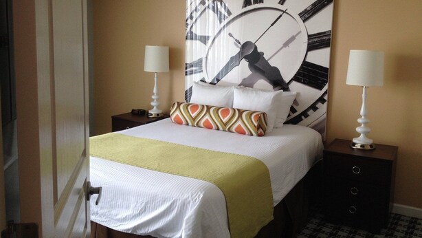 A hotel room with a large wall clock, a bed with a patterned pillow, two bedside tables with lamps, and a door. - Peak July Escape! Spacious 2BR Suite in the Heart of Victoria (Victoria)