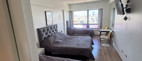Quarto de hotel com três camas, uma televisão presa à parede e uma janela com vista para a cidade.