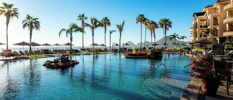 A clear blue pool with lounge chairs and palm trees, surrounded by a resort building.