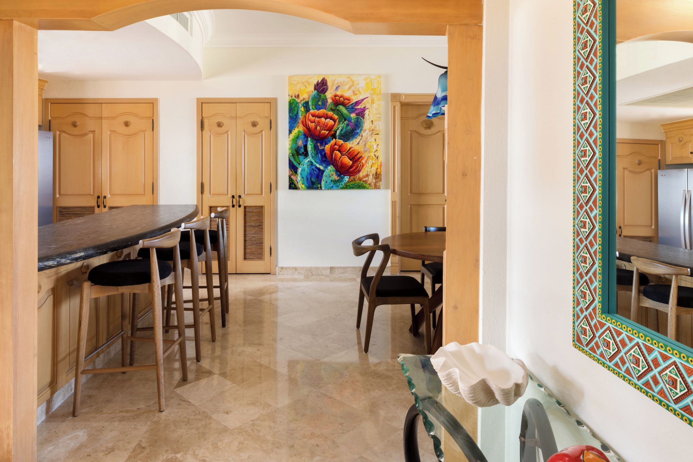 Une cuisine avec des meubles de rangement en bois, une table à manger et des chaises. Un tableau coloré orne le mur.