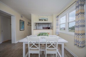 A dining area with a white table and chairs, a small kitchen area above, and a window with patterned curtains. - 5621 - Dolphin Cove Oceanfront 2BR condo with pool, deck access, and direct beach views. (Nags Head)