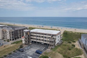 A beachfront property with a parking lot, a building with multiple floors, and a beach in the background. - 5621 - Dolphin Cove Oceanfront 2BR condo with pool, deck access, and direct beach views. (Nags Head)