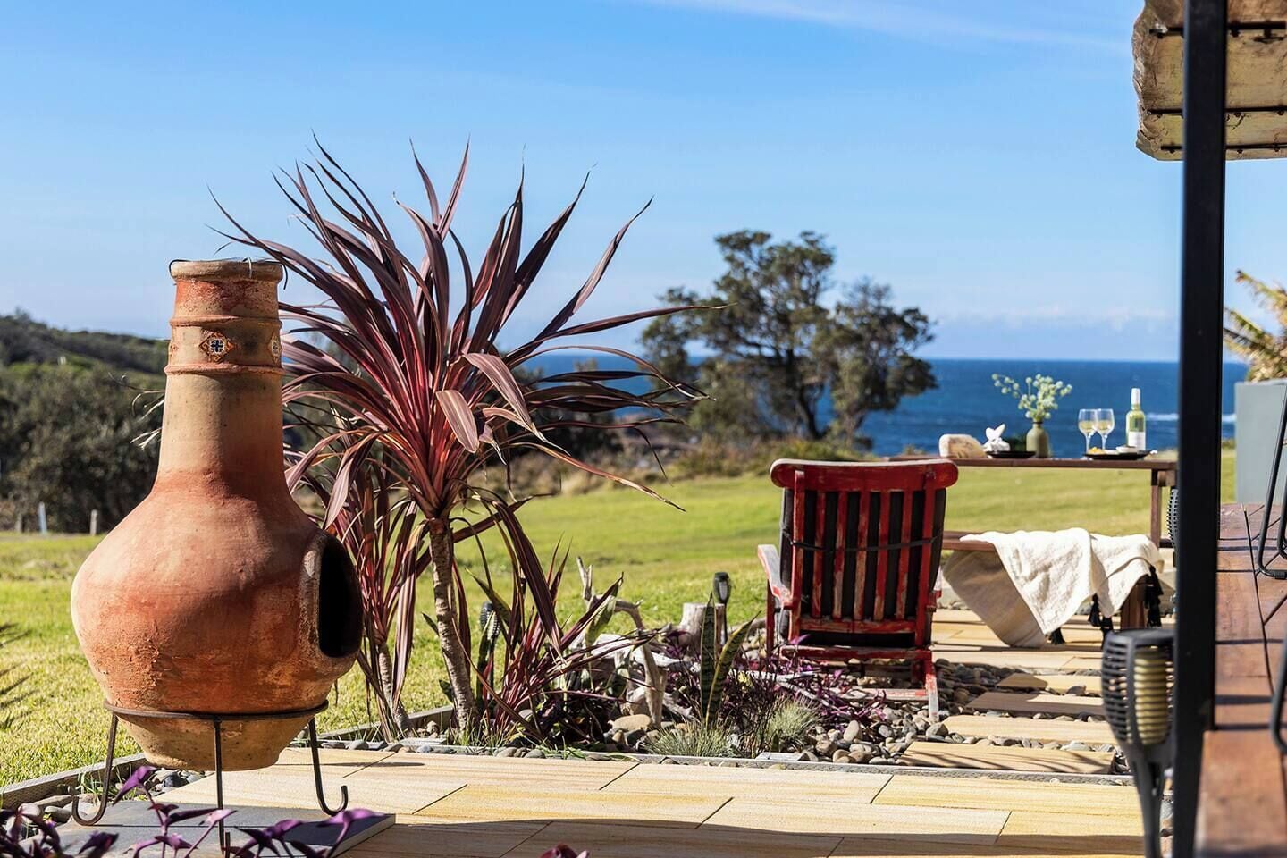 A rustic outdoor setting with a terracotta pot, a red chair, and a table with a bottle and glass, overlooking a grassy area and the sea.
