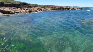 Paisagem costeira com águas claras, linha costeira rochosa e edifícios ao longe.