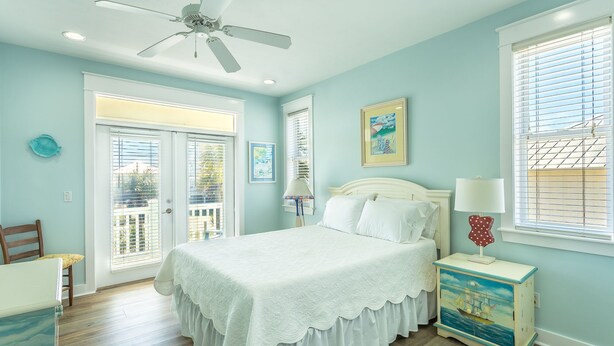 Une chambre à coucher comprenant un lit, une table de chevet, un ventilateur de plafond, une fenêtre avec des stores et une porte donnant sur un espace extérieur.