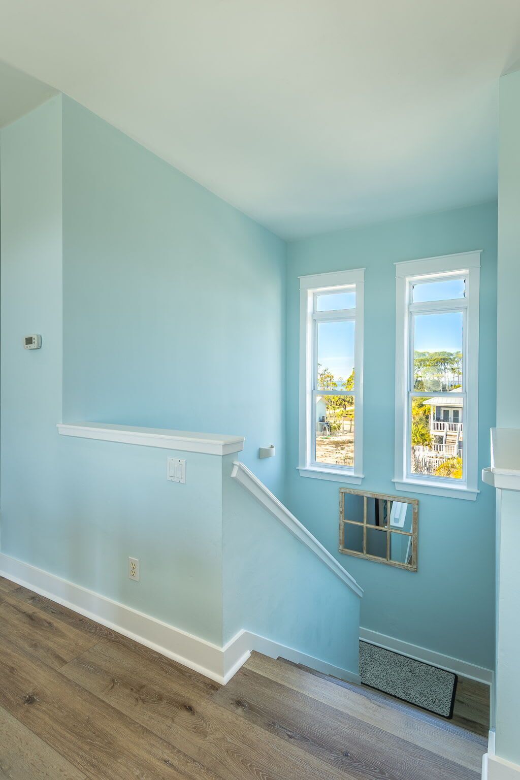 Una habitación con paredes azul claro, piso de madera y ventana que da a una casa y árboles.