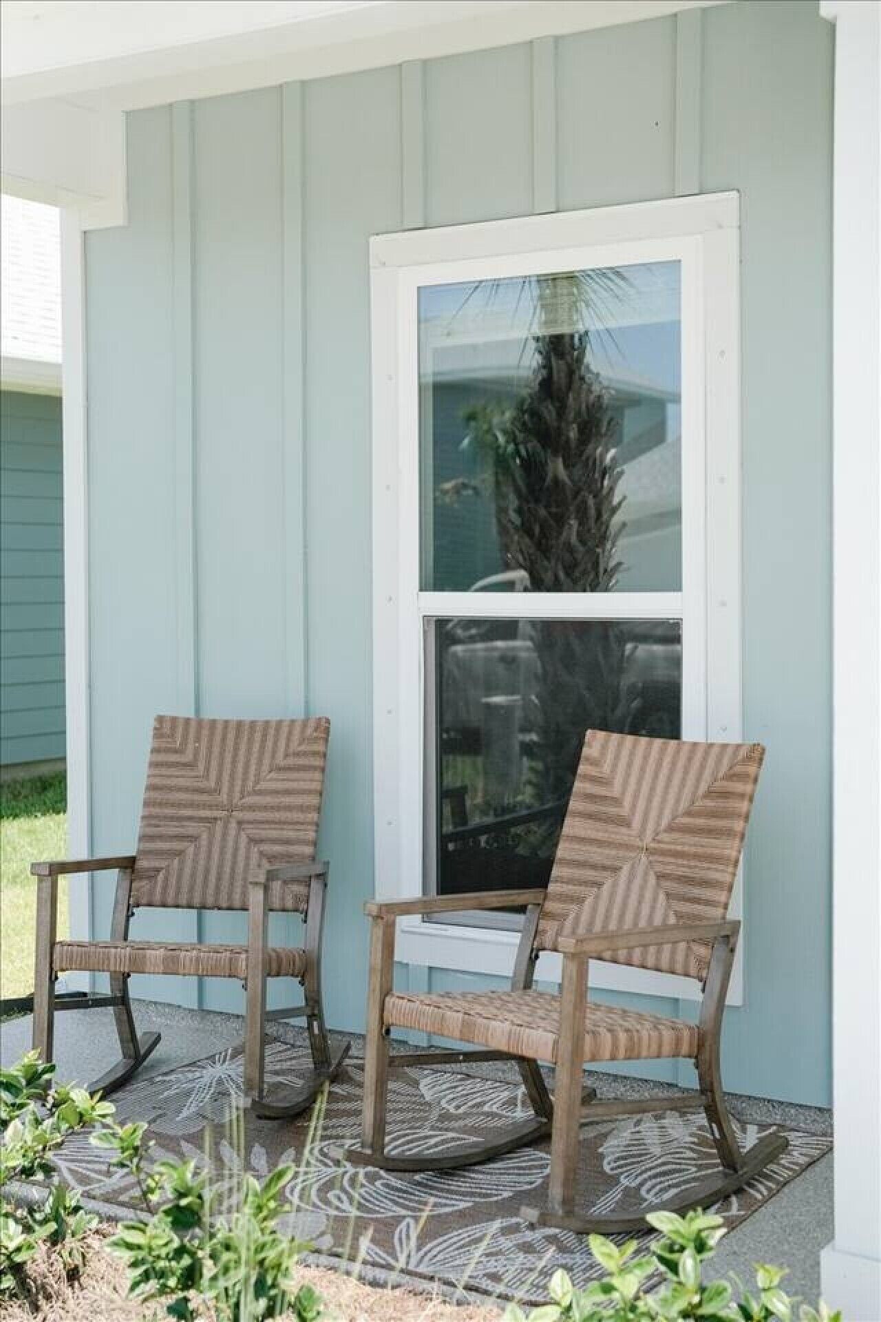 Zwei Holzstühle mit gewobener Sitzfläche und Rückenlehne stehen auf einer gemusterten Teppichmatte vor einem hellblauen Haus mit weißem Fensterrahmen.