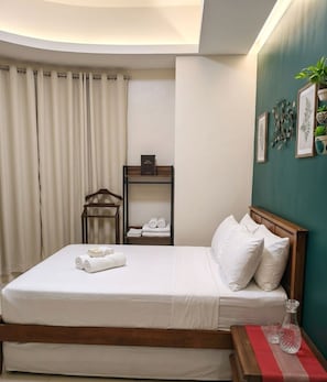 A neatly made bed with white linens and pillows, a bedside table with a glass and a red cloth, a wooden nightstand, a shelf with folded towels, and a wall with framed artwork. - Perfect Airport Suite walk to Naia 3 (Pasay City)