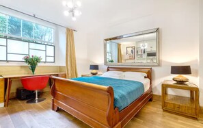 Ein Schlafzimmer mit einem hölzernen Bett, einem Schreibtisch, einem roten Stuhl, einem Fenster mit Vorhängen, einem Spiegel und einem Nachttisch mit Lampe.